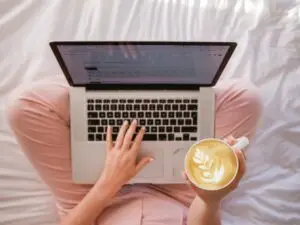 Vrouw in kleermakerszit met laptop op schoot en een kop cappuccino in haar hand